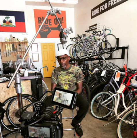 Man sitting in bicycle shop surrounded by bikes and filming equipment.